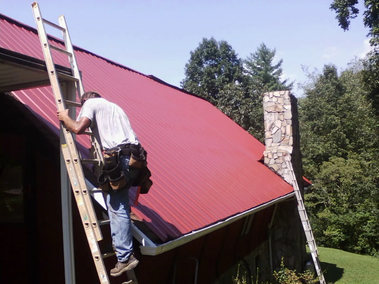 Roofer on a ladder beside a red metal roof and stone chimney — Soffit & Fascia Repair by Roofing Brothers in Claymont
